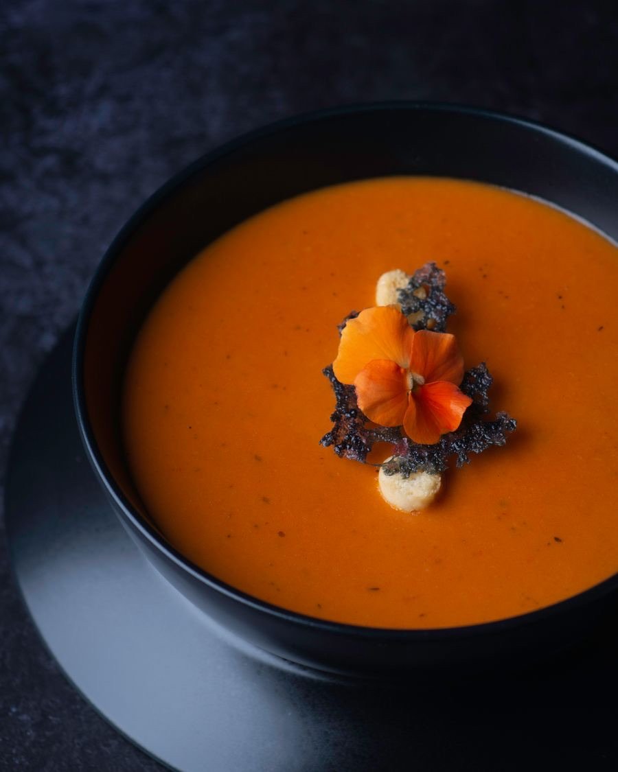 Crema anaranjada en un bol oscuro, coronada con una flor y un crujiente.